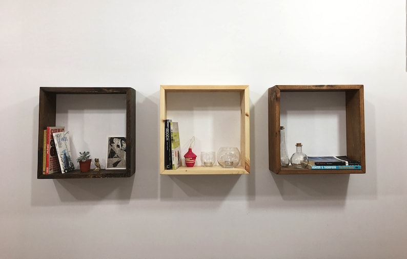 May include: Three square wooden wall shelves mounted in a horizontal row. The two outer shelves are finished in a dark brown stain, while the center shelf is a natural light wood tone. Each shelf contains various books, small plants, and glass items.
