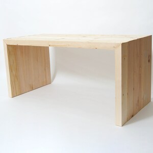 May include: A light-colored wooden desk featuring a rectangular top and two solid side panels. The desk is constructed from vertical planks of natural wood with visible grain patterns, creating a continuous U-shaped frame design.