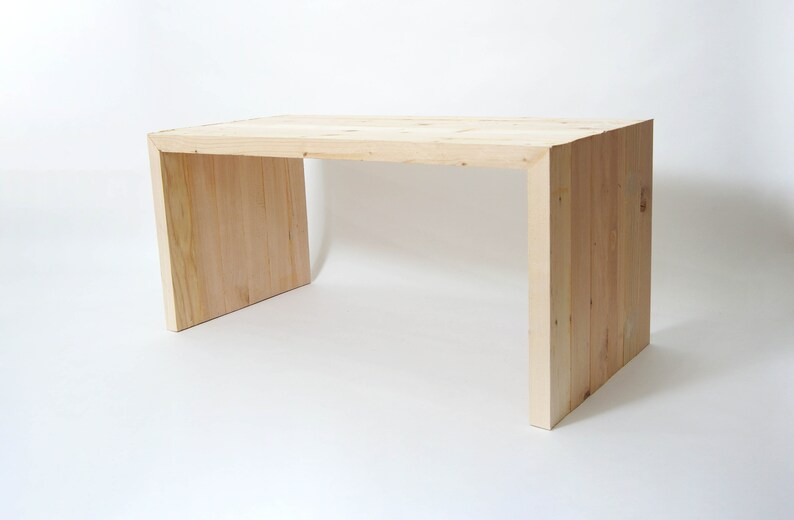 May include: A light-colored wooden desk featuring a rectangular top and two solid side panels. The desk is constructed from vertical planks of natural wood with visible grain patterns, creating a continuous U-shaped frame design.