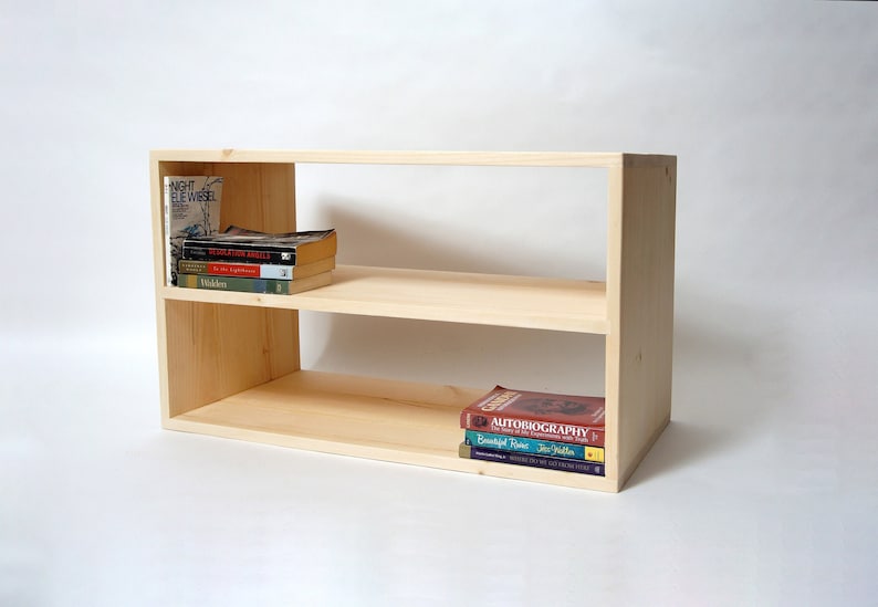 May include: A light-colored wooden bookshelf featuring two open horizontal shelves. The top shelf holds a stack of four books, including Night by Elie Wiesel and Walden. The bottom shelf holds a stack of three books, including an Autobiography.