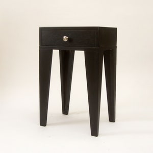 May include: A black nightstand with a single drawer and a silver knob. The nightstand has four tapered legs. The wood grain is visible on the drawer and the top of the nightstand. The nightstand is a modern design.