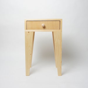 May include: A small, light-coloured wooden side table with a single drawer and a round knob. The table has four angled legs and is approximately 61 cm tall.