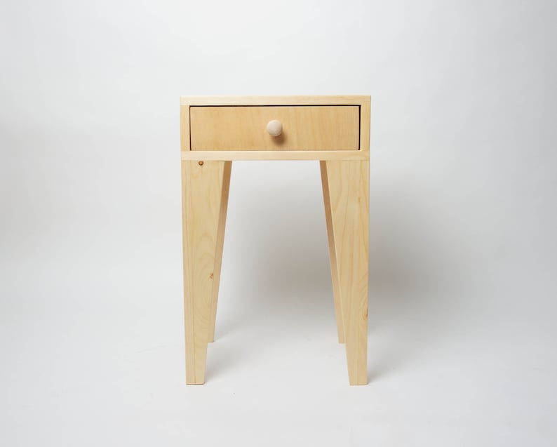 May include: A small, light-coloured wooden side table with a single drawer and a round knob. The table has four angled legs and is approximately 61 cm tall.