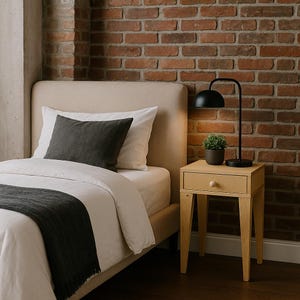 May include: A bedroom setting with a beige upholstered bed, white bedding, and a dark grey throw. A small wooden bedside table with a drawer and a black lamp with a curved arm are beside the bed. A small potted plant adds a touch of greenery.
