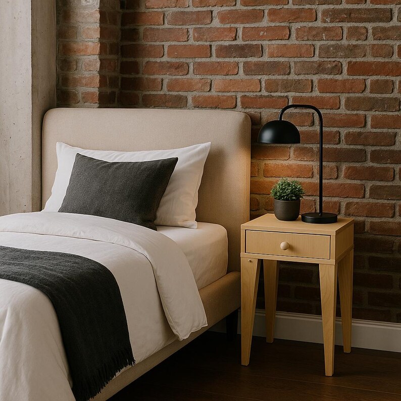 May include: A bedroom setting with a beige upholstered bed, white bedding, and a dark grey throw. A small wooden bedside table with a drawer and a black lamp with a curved arm are beside the bed. A small potted plant adds a touch of greenery.