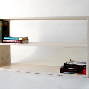 May include: A light-colored wooden bookshelf with two horizontal tiers. The top shelf holds a vertical row of books on the left and a horizontal stack of two books. The bottom shelf holds a stack of three books on the right side.