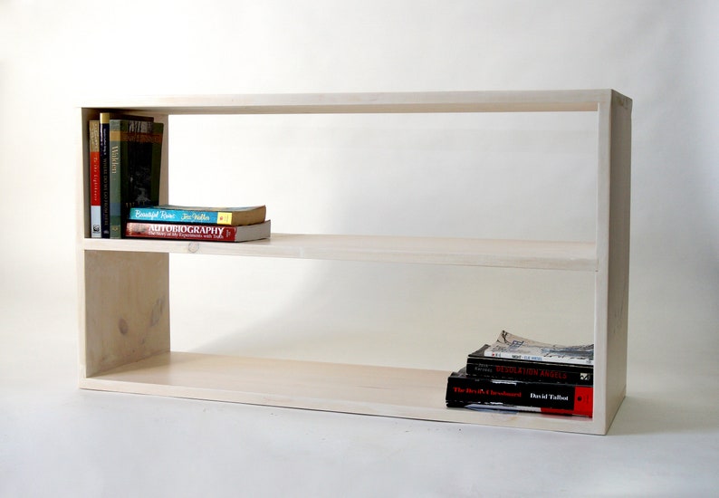 May include: A light-colored wooden bookshelf with two horizontal tiers. The top shelf holds a vertical row of books on the left and a horizontal stack of two books. The bottom shelf holds a stack of three books on the right side.