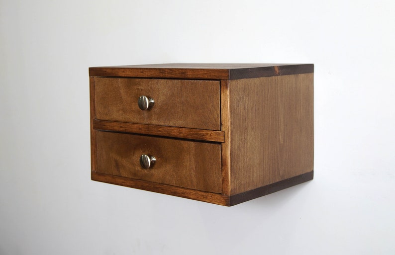 May include: A wall-mounted wooden storage unit featuring two stacked drawers with a medium brown stained finish and small, round, brushed silver metal knobs. The rectangular cabinet has a clean, simple design with visible wood grain texture.