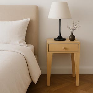 May include: A light wood bedside table with a single drawer and a round knob. A black lamp with a white lampshade sits on top, along with a small brown vase holding sprigs. A bed with a beige headboard and white bedding is partially visible.