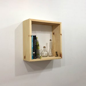 May include: A square, light-colored natural wood wall shelf featuring a simple frame construction. Inside the shelf are three books, two clear glass bottles with cork stoppers, and a small brown figurine shaped like an owl, all arranged on the base.