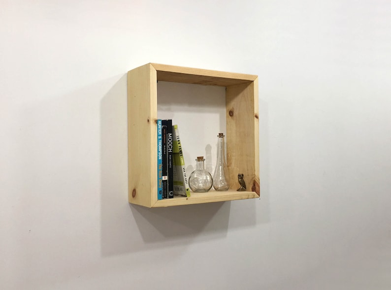May include: A square, light-colored natural wood wall shelf featuring a simple frame construction. Inside the shelf are three books, two clear glass bottles with cork stoppers, and a small brown figurine shaped like an owl, all arranged on the base.