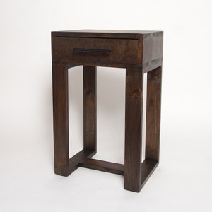 May include: A tall, rectangular side table crafted from dark brown stained wood. The table features a single drawer at the top with a thin, horizontal black metal handle. It stands on four straight legs connected by a base frame at the bottom.