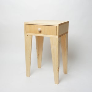 May include: A small, light-coloured wooden side table with a single drawer and a round knob. The table has four angled legs and is approximately 61 cm tall. The drawer is centered on the front of the table.