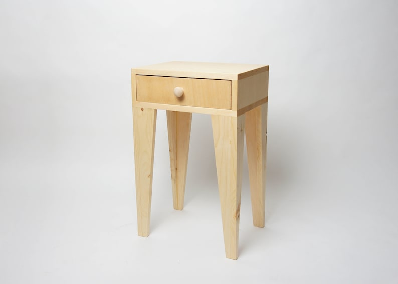 May include: A small, light-coloured wooden side table with a single drawer and a round knob. The table has four angled legs and is approximately 61 cm tall. The drawer is centered on the front of the table.