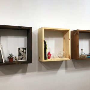 May include: Three square wall-mounted wooden shelves in varying finishes: one dark brown, one light natural wood, and one medium brown. Each shelf holds small decorative items including books, a small potted succulent, and glass containers.