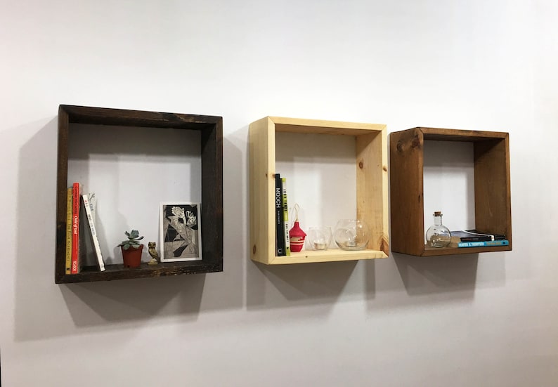 May include: Three square wall-mounted wooden shelves in varying finishes: one dark brown, one light natural wood, and one medium brown. Each shelf holds small decorative items including books, a small potted succulent, and glass containers.