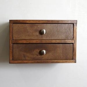 May include: A rectangular wooden storage unit featuring two stacked drawers. The unit is constructed from dark brown stained wood with a visible grain. Each drawer is equipped with a single, centered, brushed silver-toned metal knob handle.