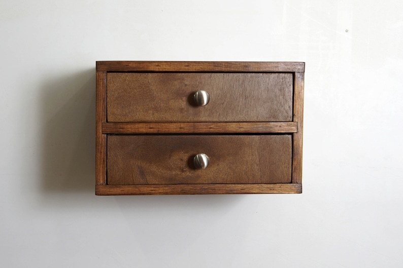 May include: A rectangular wooden storage unit featuring two stacked drawers. The unit is constructed from dark brown stained wood with a visible grain. Each drawer is equipped with a single, centered, brushed silver-toned metal knob handle.