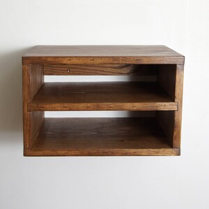 May include: A rectangular wall-mounted wooden shelf unit featuring a dark brown stained finish. The unit consists of a solid top surface and one horizontal middle shelf, creating two open storage compartments. Two small metal screw heads are visible.