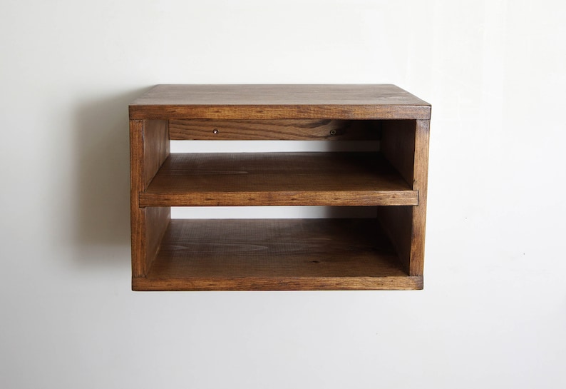 May include: A rectangular wall-mounted wooden shelf unit featuring a dark brown stained finish. The unit consists of a solid top surface and one horizontal middle shelf, creating two open storage compartments. Two small metal screw heads are visible.