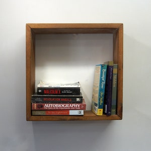 May include: A square wooden wall shelf containing a stack of five books lying horizontally and three books standing vertically. The titles include Night, Malcolm X, Desolation Angels, Autobiography, To the Lighthouse, Beautiful Ruins, and Walden.