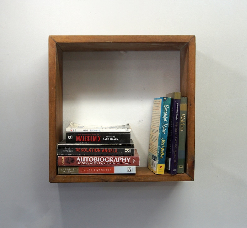 May include: A square wooden wall shelf containing a stack of five books lying horizontally and three books standing vertically. The titles include Night, Malcolm X, Desolation Angels, Autobiography, To the Lighthouse, Beautiful Ruins, and Walden.