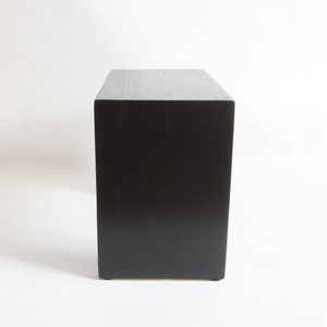 May include: A rectangular wooden block with a dark black finish and a visible wood grain texture. The object is oriented vertically, showcasing a flat top surface and a solid, smooth front panel with clean, sharp edges on all sides.