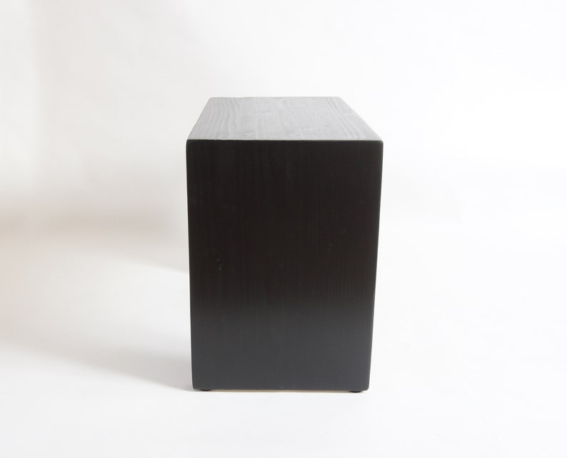 May include: A rectangular wooden block with a dark black finish and a visible wood grain texture. The object is oriented vertically, showcasing a flat top surface and a solid, smooth front panel with clean, sharp edges on all sides.