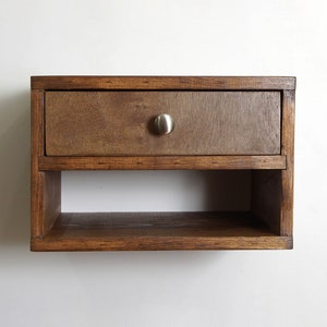 May include: A wall-mounted wooden shelf featuring a single rectangular drawer with a brushed metal knob. The unit is constructed from dark stained wood and includes an open storage compartment located directly beneath the drawer section.