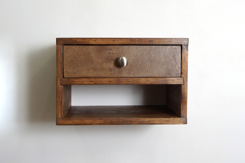May include: A wall-mounted wooden shelf featuring a single rectangular drawer with a brushed metal knob. The unit is constructed from dark stained wood and includes an open storage compartment located directly beneath the drawer section.