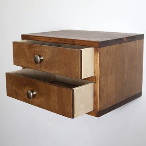 May include: A small wooden storage unit with two drawers, featuring a dark brown stained finish and light-colored interior drawer sides. Each drawer is equipped with a single, round, silver-toned metal knob centered on the front panel.
