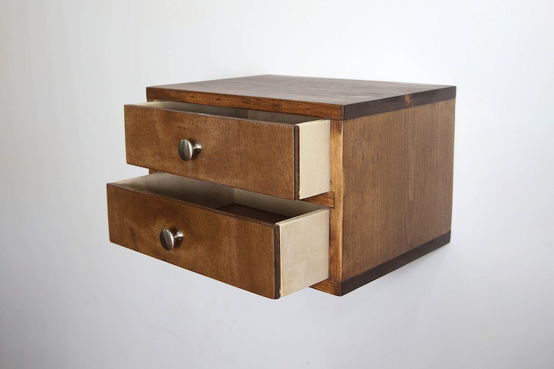 May include: A small wooden storage unit with two drawers, featuring a dark brown stained finish and light-colored interior drawer sides. Each drawer is equipped with a single, round, silver-toned metal knob centered on the front panel.