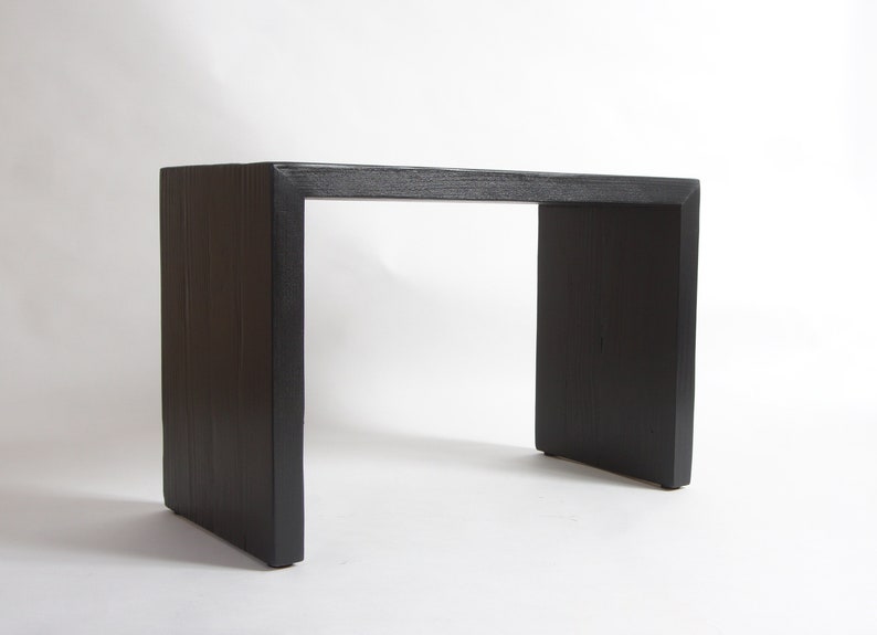 May include: A black console table with a rectangular top and two thick, solid vertical side supports. The piece features a visible wood grain texture throughout its surface, constructed in a simple, continuous U-shaped frame design.
