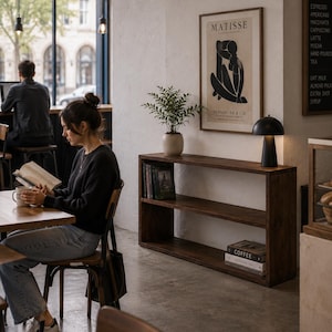May include: A dark wood console table with two open shelves, featuring a potted green plant, a black dome-shaped table lamp, and stacked books. A framed Matisse print hangs on the white wall above the table in a coffee shop setting.
