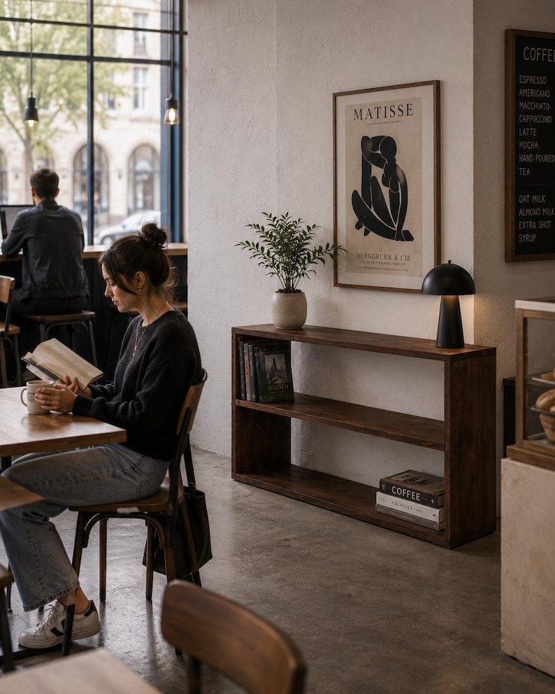 May include: A dark wood console table with two open shelves, featuring a potted green plant, a black dome-shaped table lamp, and stacked books. A framed Matisse print hangs on the white wall above the table in a coffee shop setting.