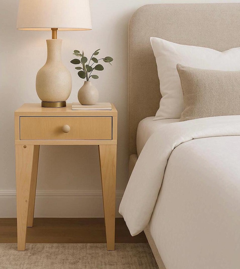 May include: A light wood bedside table with a single drawer and a round knob. A beige lamp with a white lampshade sits on top, next to a small vase with greenery. A bed with a beige headboard and white bedding is in the background.