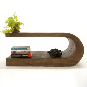 May include: A dark brown wooden side table with a curved design. The table has two shelves, one on top and one below. A potted plant, a stack of books, and a camera are on the table.
