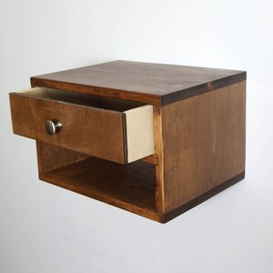 May include: A wall-mounted floating nightstand crafted from dark brown stained wood. The unit features a single pull-out drawer with a small, round, silver-toned metal knob and an open storage shelf located directly beneath the drawer compartment.