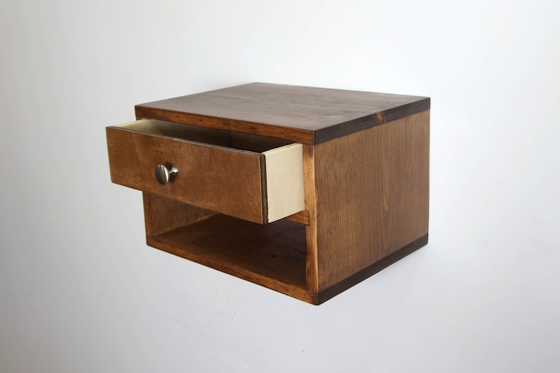 May include: A wall-mounted floating nightstand crafted from dark brown stained wood. The unit features a single pull-out drawer with a small, round, silver-toned metal knob and an open storage shelf located directly beneath the drawer compartment.
