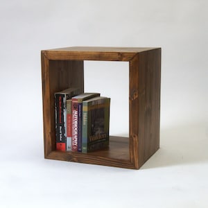 May include: A dark brown wooden cube shelf with a single open shelf. The shelf is filled with a variety of books, including "Autobiography" and "Walden".