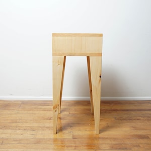 May include: A light-coloured wooden side table with a rectangular top and four angled legs. The table is made of natural wood and has a simple, minimalist design. The table's height is approximately 76 cm.