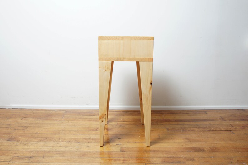 May include: A light-coloured wooden side table with a rectangular top and four angled legs. The table is made of natural wood and has a simple, minimalist design. The table's height is approximately 76 cm.