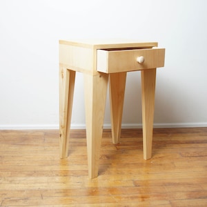 May include: A light-coloured wooden bedside table with a single drawer that is partially open. The drawer has a round wooden knob. The bedside table has four angled legs. The table is on a wooden floor.