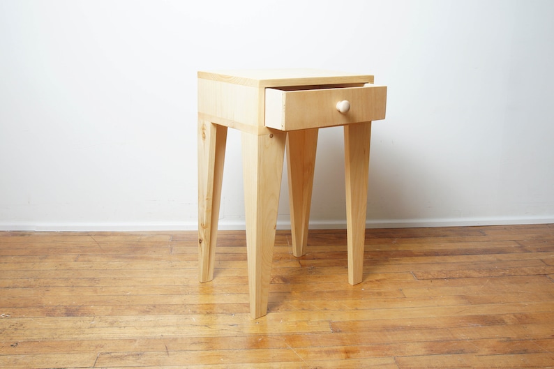May include: A light-coloured wooden bedside table with a single drawer that is partially open. The drawer has a round wooden knob. The bedside table has four angled legs. The table is on a wooden floor.