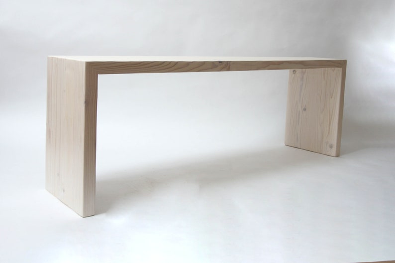 May include: A rectangular bench constructed from light-colored wood with a visible grain pattern. The furniture piece features a continuous waterfall design where the flat top surface transitions seamlessly into two vertical side support legs.