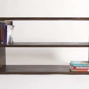 May include: A rectangular dark brown wooden bookshelf with two horizontal tiers. The top shelf contains three books standing upright on the left side. The bottom shelf holds a stack of three books lying flat on the right side of the surface.