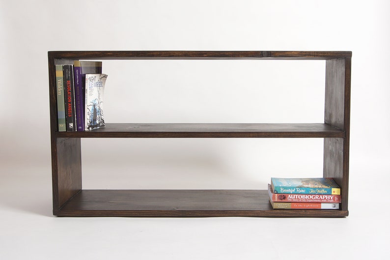May include: A rectangular dark brown wooden bookshelf with two horizontal tiers. The top shelf contains three books standing upright on the left side. The bottom shelf holds a stack of three books lying flat on the right side of the surface.