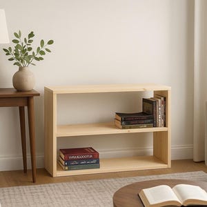 May include: A low, rectangular, light-colored wooden bookshelf with two open shelves. The top shelf holds a small collection of books standing upright and stacked horizontally. The bottom shelf contains two books stacked horizontally on the left side.