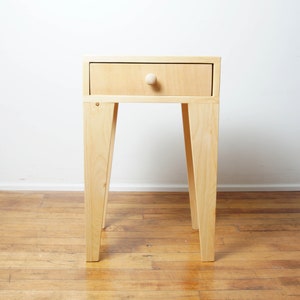 May include: A light-coloured wooden side table with a single drawer and a round knob. The table has four angled legs and is set against a white wall and wooden floor.