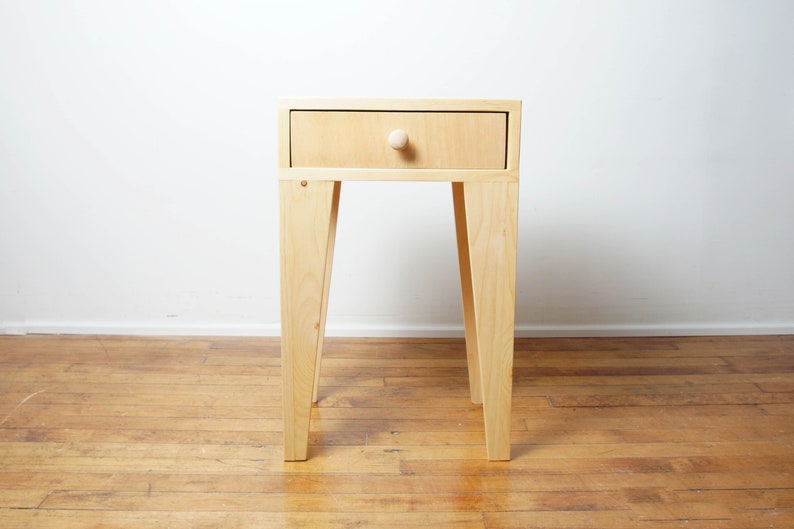 May include: A light-coloured wooden side table with a single drawer and a round knob. The table has four angled legs and is set against a white wall and wooden floor.